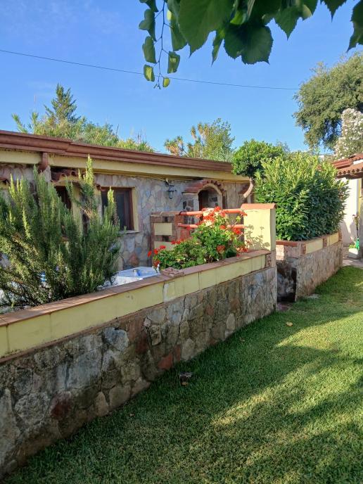 Maison en pierre avec jardin bien entretenu, parterres de fleurs et mur rustique dans un environnement verdoyant et ensoleillé.