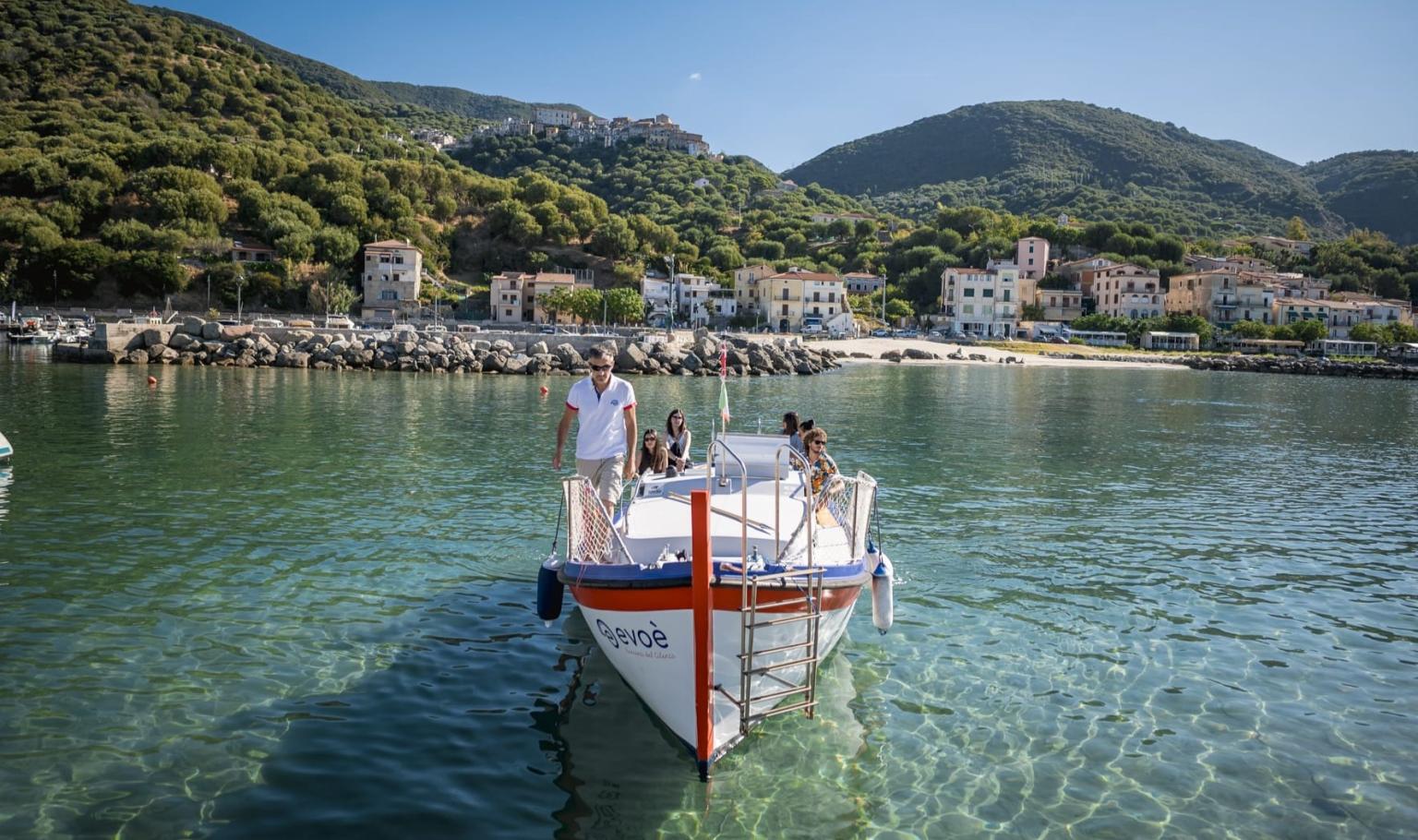 Bateau touristique naviguant dans des eaux cristallines, avec en arrière-plan un village côtier pittoresque entouré de collines verdoyantes.