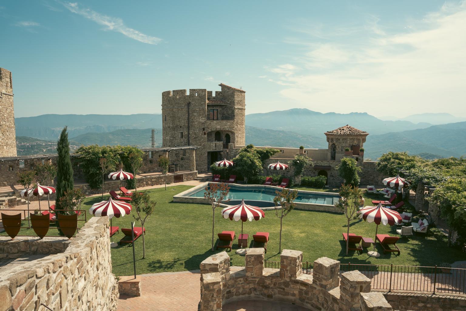 Château en pierre avec piscine panoramique, jardin soigneusement entretenu et parasols rouges, niché au cœur des montagnes et entouré de paysages à couper le souffle.