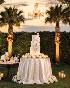 Gâteau de mariage blanc à trois étages, orné de roses, de bougies et d’un lustre, dans un élégant décor de mariage en plein air au coucher du soleil.
