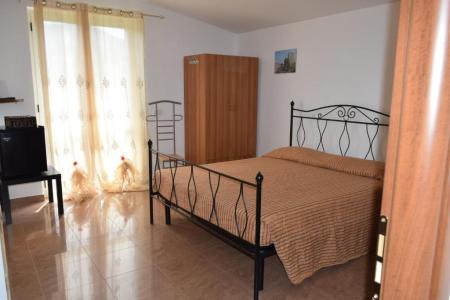 Bright bedroom with a wrought-iron double bed, wooden wardrobe, ceramic tile flooring and large windows with light-colored curtains.