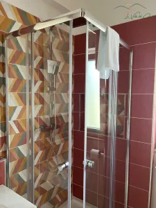 Moderne Duschkabine mit transparenten Glaswänden, verchromten Armaturen und farbenfrohen geometrischen Fliesen in Terrakotta, Beige und Salbeigrün.