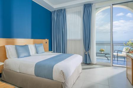 Camera d’hotel vista mare con letto matrimoniale, arredi azzurri e balcone panoramico sul mare.