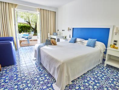 Bright and elegant Mediterranean-style double bedroom, featuring a blue headboard, decorative majolica tile flooring, white furnishings, and beige curtains.