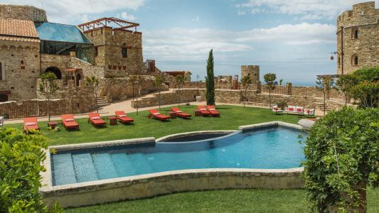 Piscine extérieure nichée dans un charmant château en pierre, avec un solarium aménagé de transats rouges et une pelouse verdoyante.