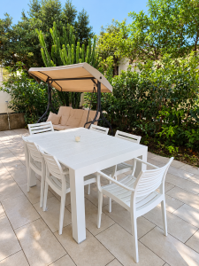 White garden furniture with table and chairs, swing and green plants on an outdoor terrace.