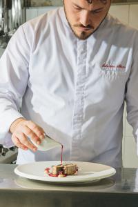 Chef wearing a white jacket plating a gourmet dish with red sauce in a professional kitchen.